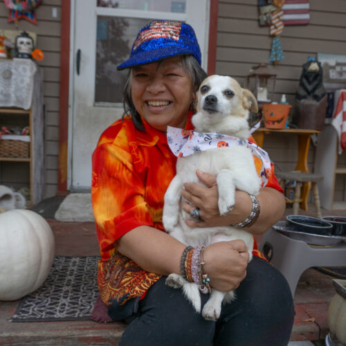 A woman in a red shirt sits on her porch and holds a small dog.