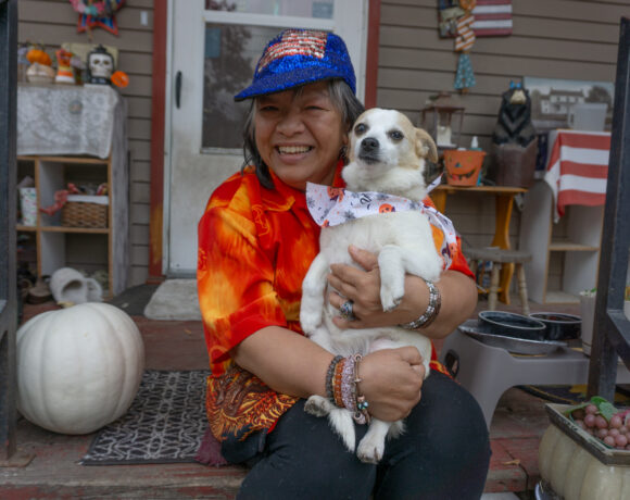 A woman in a red shirt sits on her porch and holds a small dog.