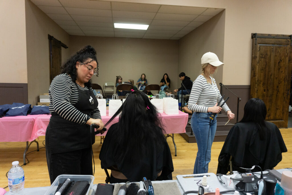 Two women in striped shirts straighten the hair of two women, as seen from the back.