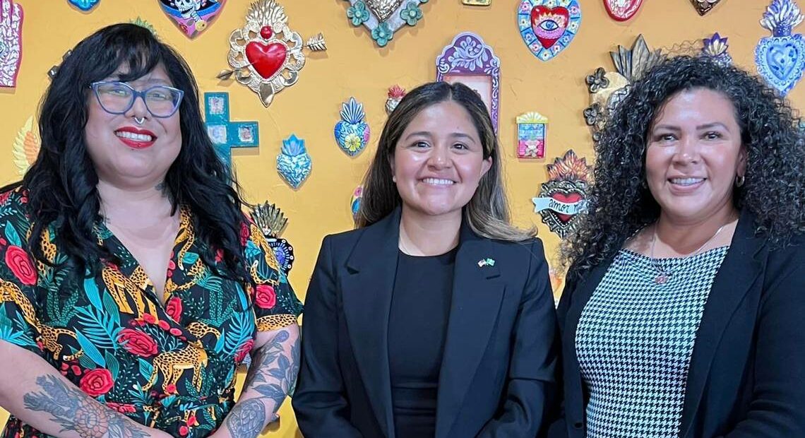 Three women stand in front of a yellow wall, posing for a group photo.