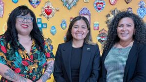 Three women stand in front of a yellow wall, posing for a group photo.