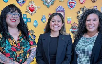 Three women stand in front of a yellow wall, posing for a group photo.