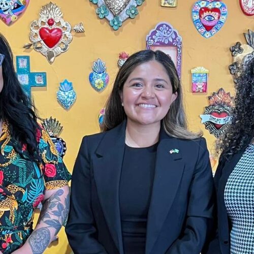 Three women stand in front of a yellow wall, posing for a group photo.