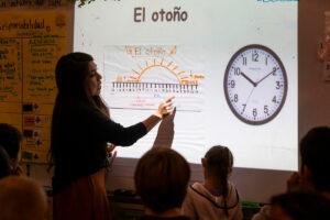 A woman stands at projector screen. An image of a clock and words in Spanish are projected on the screen.