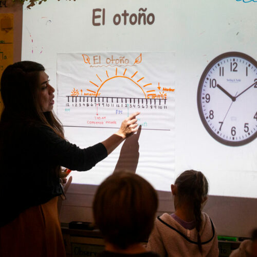 A woman stands at projector screen. An image of a clock and words in Spanish are projected on the screen.