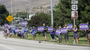 A group of people gather on the side of the road. They are holding purple signs that read "St. Joseph nurses deserve a fair contract."