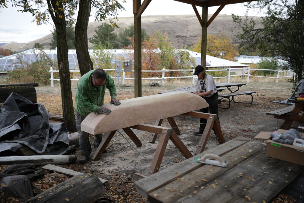 An adult and a middle schooler sand a dugout canoe. They are outside in the middle of autumn.