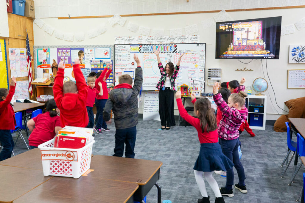 A woman teaches a class, with her hands raised above her head. The students in her class are gathered around her, and copy her movement.