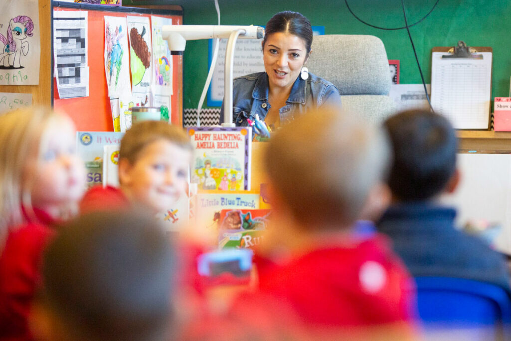 A woman speaks, sitting from a desk in a classroom. A group of children are seated in front of her.