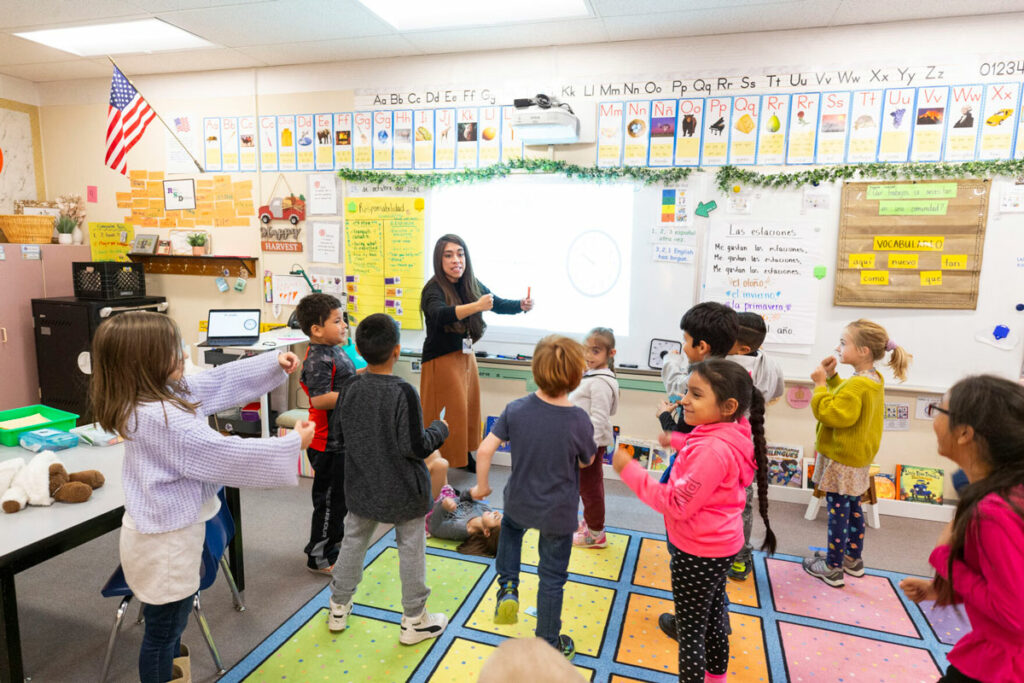 A woman is teaching in front of a classroom, her arms stretched in front of her. Her students copy her movements.