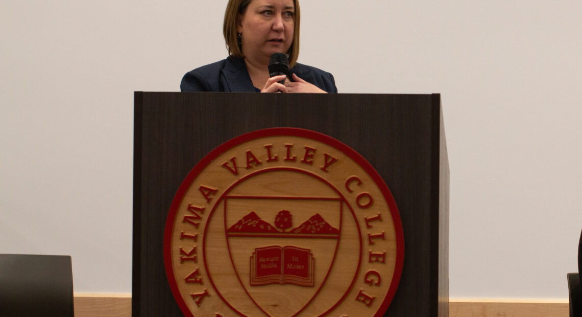 A woman stands behind a podium. She is speaking, and holding a microphone.