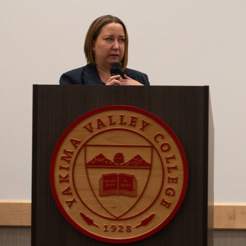 A woman stands behind a podium. She is speaking, and holding a microphone.