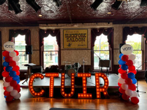 A stage is framed by red, white and blue balloon towers. in the middle of the stage is a red, lighted CTUIR sign. The stage is on a wooden floor with windows all around it.