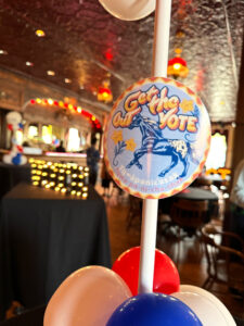 A blue and orange "get out the vote" sticker is on a white tower of red, white and blue balloons. The background of the photo is blurry, but there is a lighted 'vote' sign in the distance.