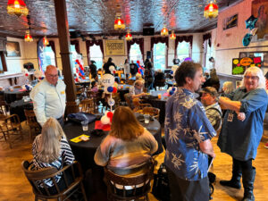 A large group of people stand in a room with wooden floors. Tall windows surround the entire room.