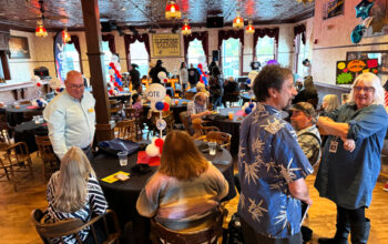 A large group of people stand in a room with wooden floors. Tall windows surround the entire room.