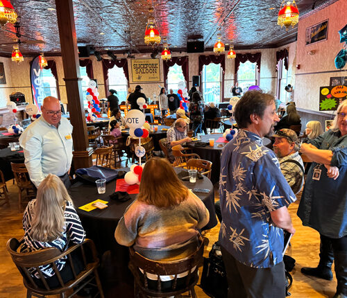 A large group of people stand in a room with wooden floors. Tall windows surround the entire room.