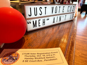 A sign that reads "Just vote cuz 'meh' ain't it' sits on a wooden bar top with a red balloon and a flyer about the voter registration drive.