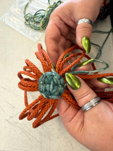 A woman with bright green fingernails is holding yarn cords that are orange and are wrapped with turquoise yarn in the center.