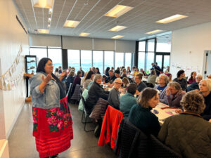 A woman in a red floral skirt and jean jacket stands in front of a crowd of people who are all gathered at long white tables.