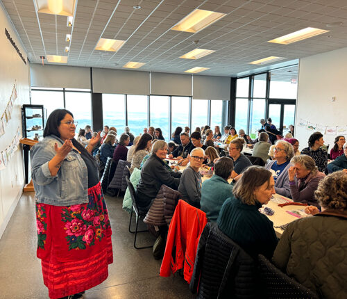 A woman in a red floral skirt and jean jacket stands in front of a crowd of people who are all gathered at long white tables.