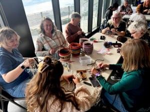 Women is all around a white table. There are colorful woven baskets on top of the table. All of the women are working on weaving projects. They have cords in their hands and are looking down.