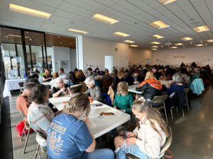 A white room is filled with tables full of people, who are all seated at white folding tables.