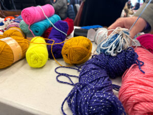 A white table is covered in multi colored balls of yard. Someone's hands are holding some white yarn.