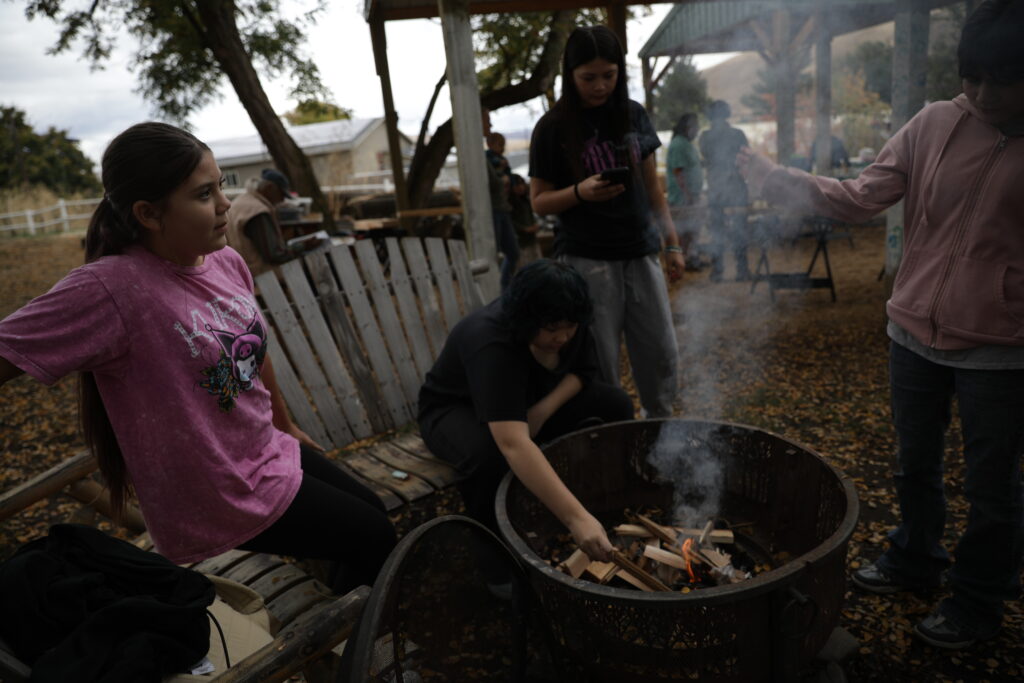 A group of kids gather around a fire pit. Two of them sit on a bench. Two of them are standing next to the fire. Smoke billows up from the fire.