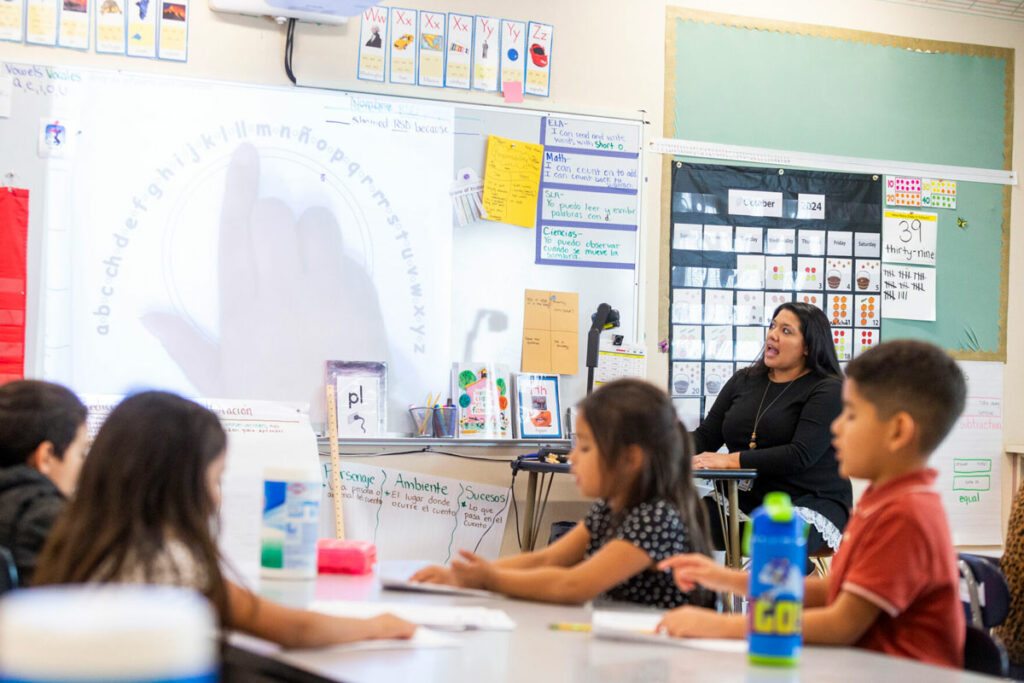 A woman is teaching in front of a document camera in a classroom. Her students watch the lesson.