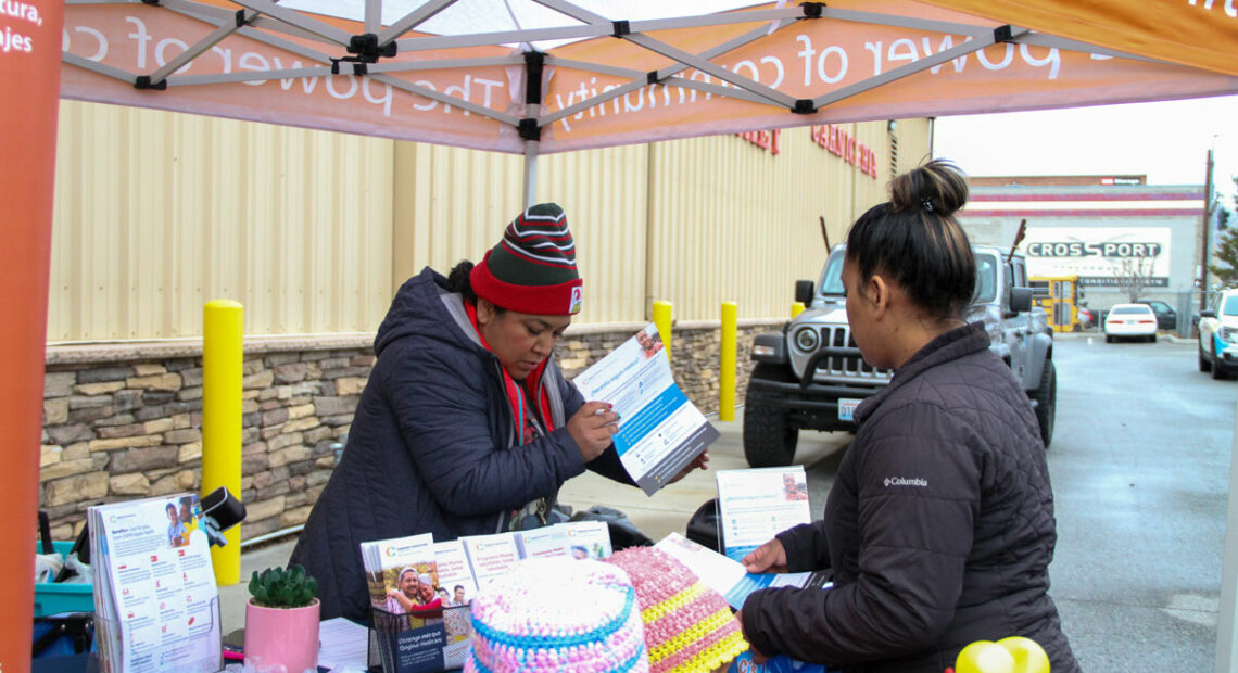 A woman in a beanie holds a packet of paper. She talks to a woman in a black puffer jacket.