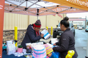 A woman in a beanie holds a packet of paper. She talks to a woman in a black puffer jacket.