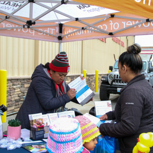 A woman in a beanie holds a packet of paper. She talks to a woman in a black puffer jacket.