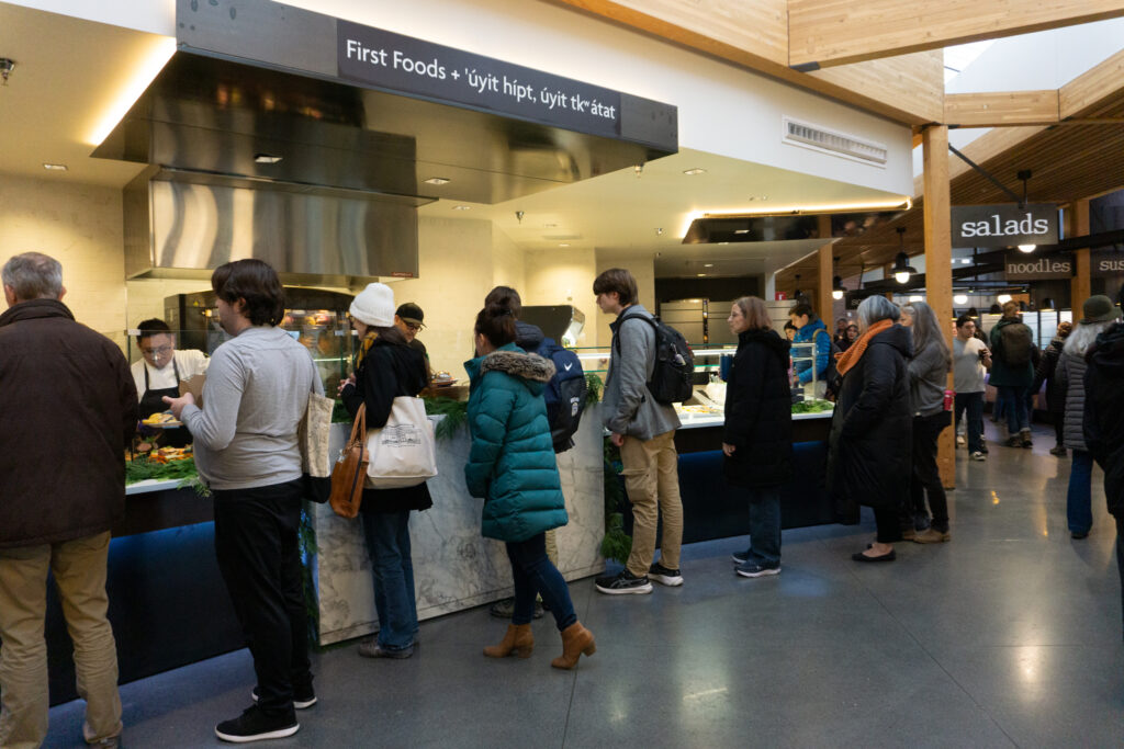 A group of people stand in a food line with a "first foods" sign above them. They are in a college cafeteria.