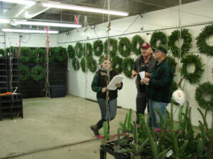 Chastagner stands against a wall of green Christmas wreaths as a woman with a clipboard walks through the facility. 