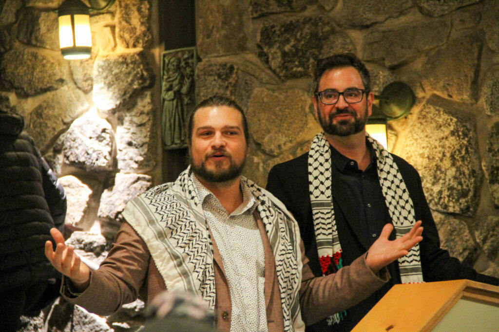 Two men are speaking. They are standing in front of a stone wall.