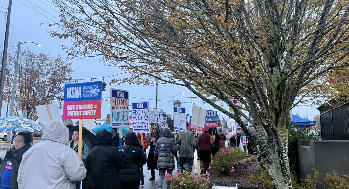 Nurses hold signs that include messages like "safe staffing = patient safety." They are walking on a sidewalk outside on a cloudy day.