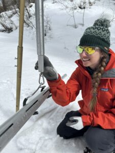 A woman in a gray beanie and a red coat with sunglasses holds big steel tools for measuring snowpack in the snow. A forest is around her.