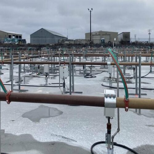 Gold pipes are attached to silver pipes that are sticking into the ground. The rows of pipes go on as far as the picture. There are buildings in the very background. The sky is gray.