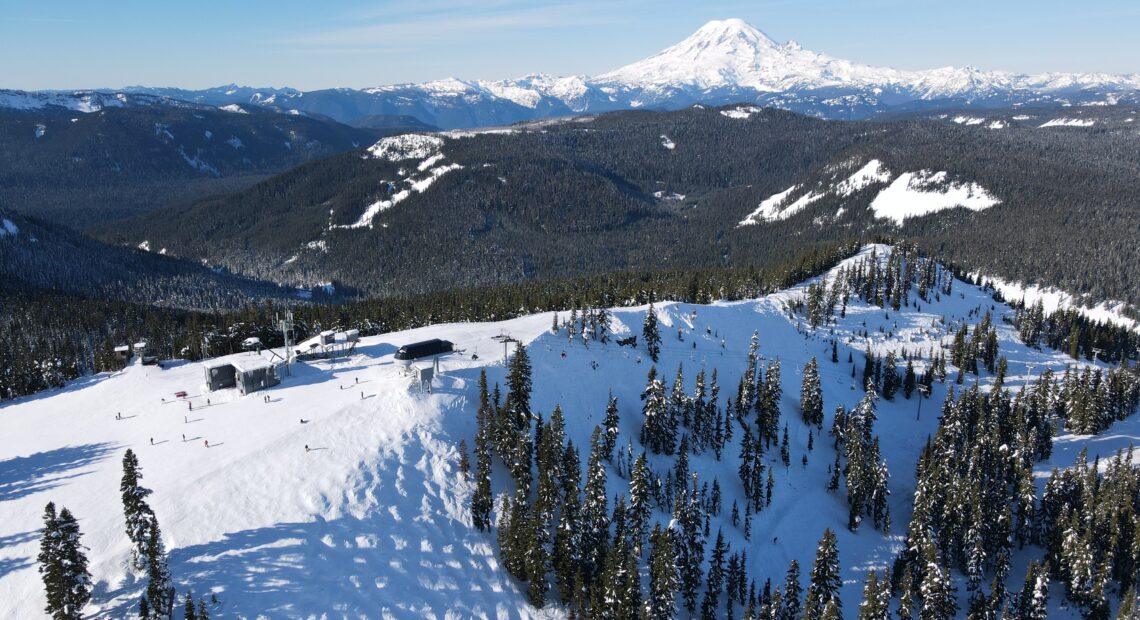 A bird's-eye view shows white ski hills flanked by evergreen trees. A snow covered mountain rises in the distance.