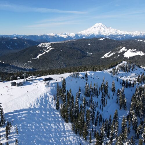 A bird's-eye view shows white ski hills flanked by evergreen trees. A snow covered mountain rises in the distance.