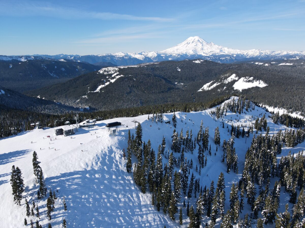 A bird's-eye view shows white ski hills flanked by evergreen trees. A snow covered mountain rises in the distance.