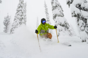 A skier in orange pants and a bright green jacket pushes through powder and snow covered trees.