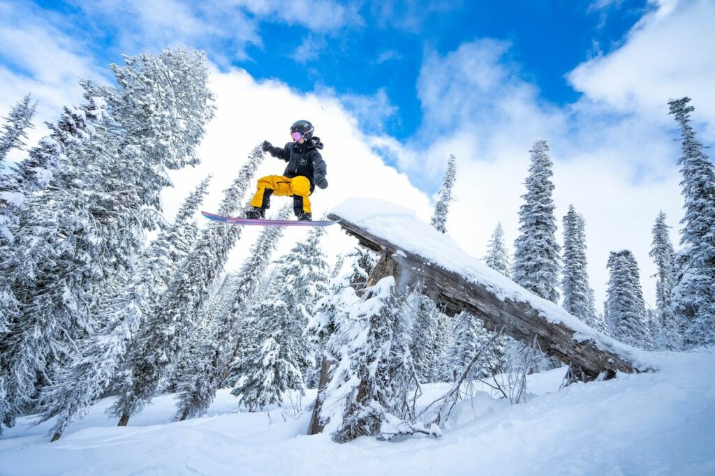 A snowboarder in a black jacket and yellow paints sails off a jump against a backdrop of snow covered trees. 