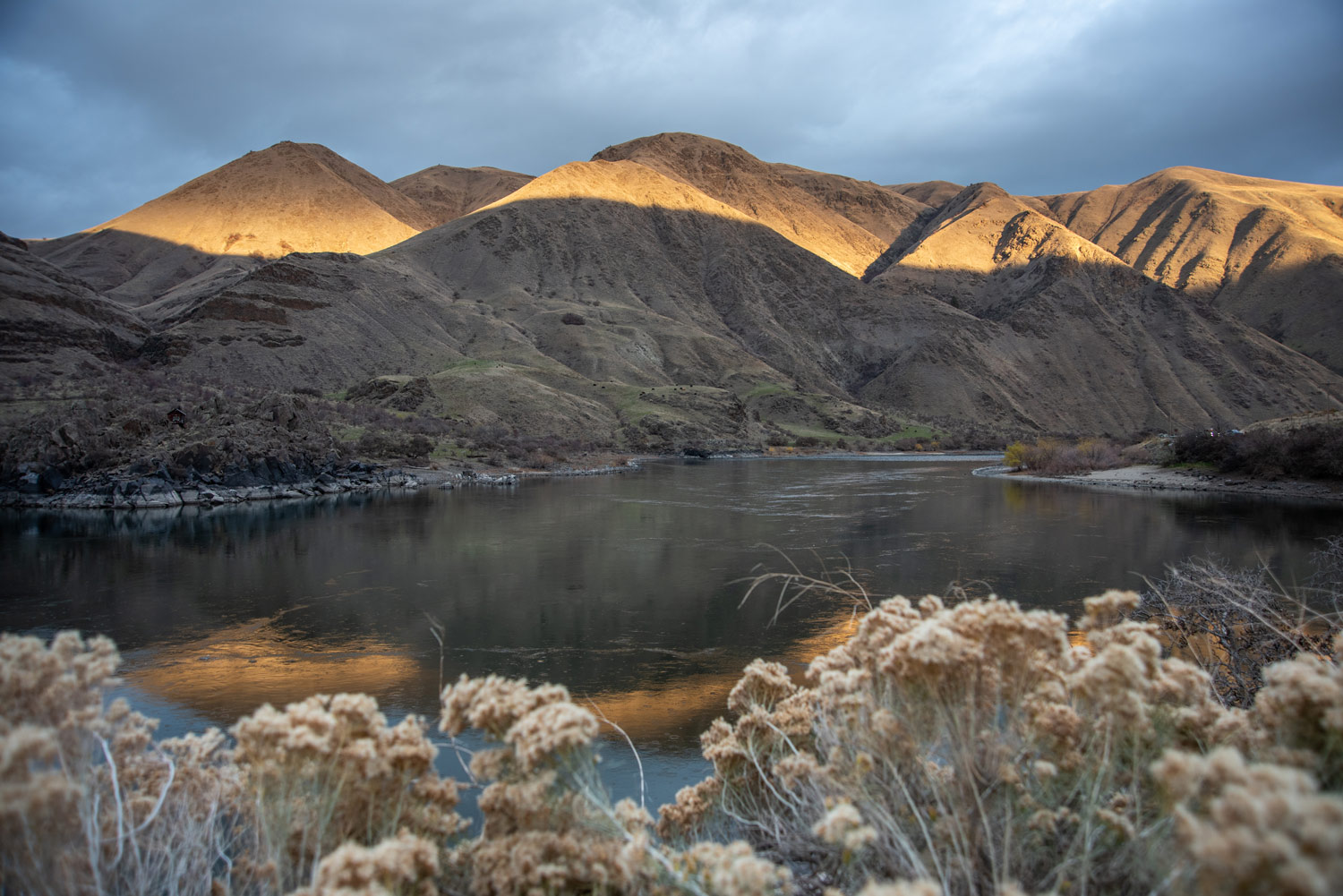 sun sets on hills in a canyon with a river in the foreground