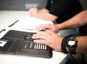 Four hands are shown on top of a white table. The hands in the foreground are on top of a notebook with a calculator.