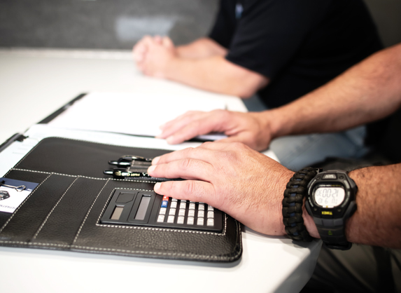 Four hands are shown on top of a white table. The hands in the foreground are on top of a notebook with a calculator.