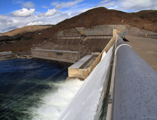 The top of a dam stretches across the photo. In the bottom of the photo, there is water, some of which is churning and white. In the back of the photo is a brown hill, blue skies and white clouds.