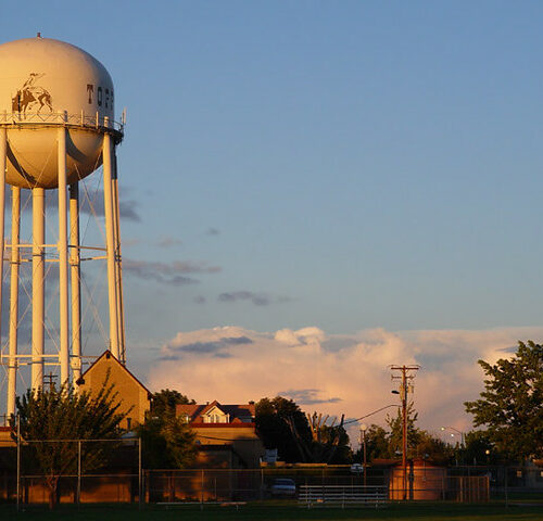 A white water tower stands tall above buildings and trees. The sky is blue with whispy white clouds.