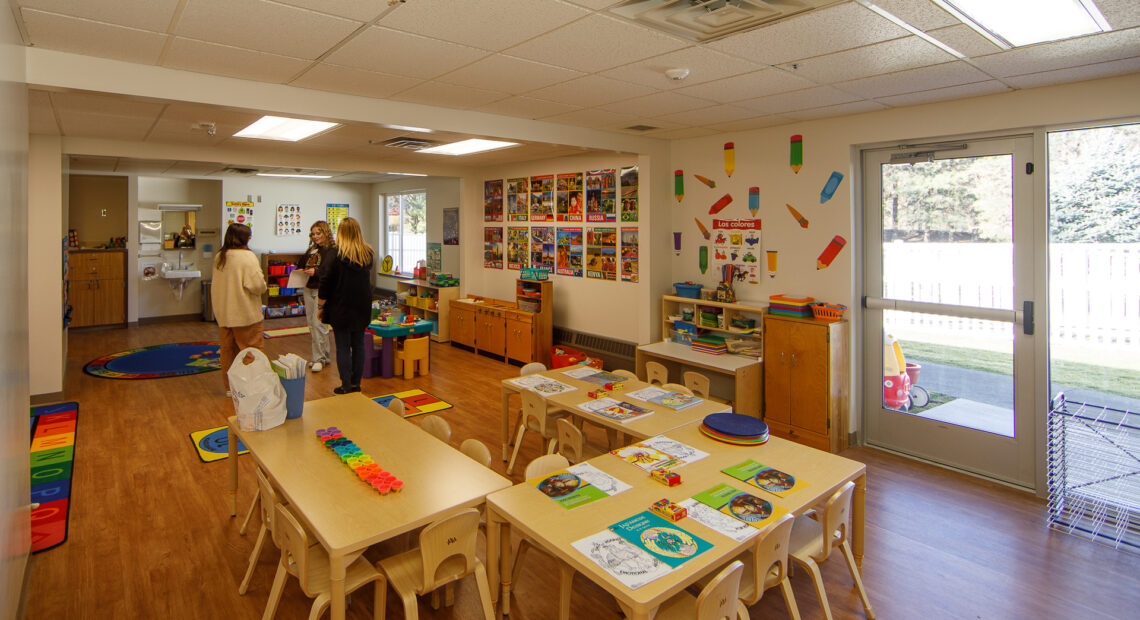 A photo shows a daycare table on hardwood floors with teal, orange and green shaped toys on top. People stand in the distance chatting.